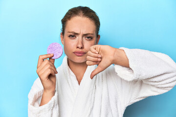 Blonde in bathrobe with facial sponge in studio showing a dislike gesture, thumbs down....