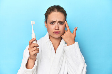 Blonde in bathrobe with electric toothbrush in studio showing a disappointment gesture with...