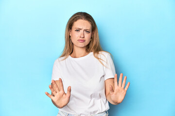 Blonde young woman in white t-shirt on blue background rejecting someone showing a gesture of...