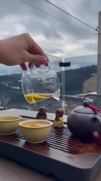 Vertical screen hand gently pours tea from glass chahai over Buddha figurines during outdoor tea ceremony small teapot two cups and chaban on table peaceful natural background