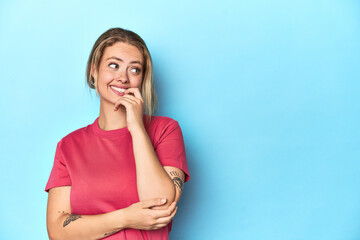 Blonde young woman in red t-shirt on blue background relaxed thinking about something looking at a copy space.