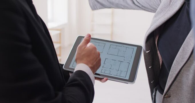 Unknown interior designer holding digital tablet with floor plan of new house with blueprint construction project, discussing with client all details, talking about house floorplan