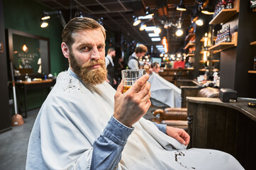 Handsome man enjoying his drink in barbershop. Male customer relaxing while getting a haircut. Confident man holding glass and looking at camera.
