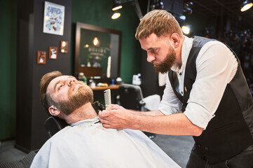 Obraz premium Professional barber trimming client's beard. Male hairdresser serving client with thin beard by clipper. Isolated photo of man getting his haircut done.