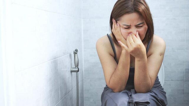 asian woman sitting on the toilet and experiencing foul-smelling stools and diarrhea, possibly caused by digestive issues such as inflammatory bowel disease, infections, food intolerances, or allergie