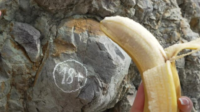 Banana Rock Symbolism: Hand peels fruit at rock, age warning at blurred seaside daytime