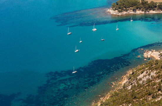 Vue a&eacute;rienne sur une baie de mer bleue turquoise transparente avec des fonds marins et des voiliers au mouillage pr&egrave;s du littoral avec des rochers et de la garrigue