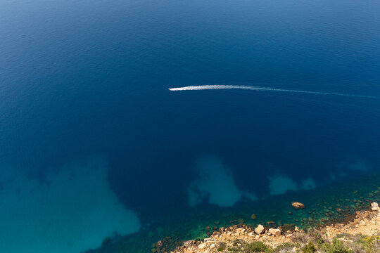 Mer de couleur bleu et turquoise transparent en vue a&eacute;rienne avec un bateau de plaisance hors-bord naviguant avec de l'&eacute;cume blanche et un bout de littoral avec des rochers et de la garrigue