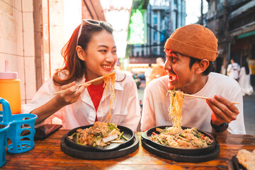 Asian couple man and woman enjoy eating street food noodle at night market. Traveler Asian couple blogger Happy tourists with Traditional food local road at thailand bangkok city.