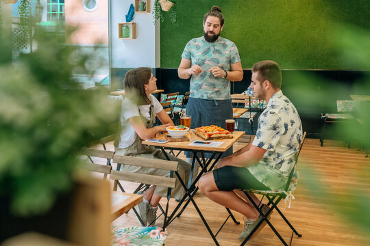 Waiter in an apron engaging with young customers seated at a table, discussing the menu while enjoying food and drinks in a modern restaurant featuring a vibrant green wall