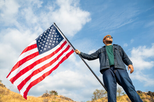 Portrait of cheerful young man looking forward while standing proudly with holding USA flag over his head against wind and blue sky, happy american holding the United states flag celebration