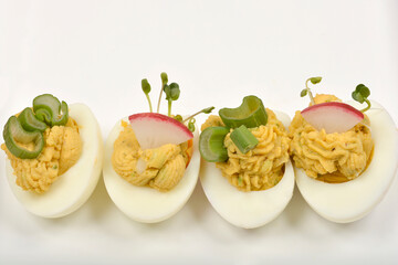 Four deviled egg halves, each uniquely garnished with celery, radish, and sprouts, are arranged on a white surface
