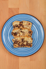 Three slices of a light-colored cake with visible pieces of chocolate, walnuts, raisins, and orange peel are arranged on a blue plate over a wooden surface