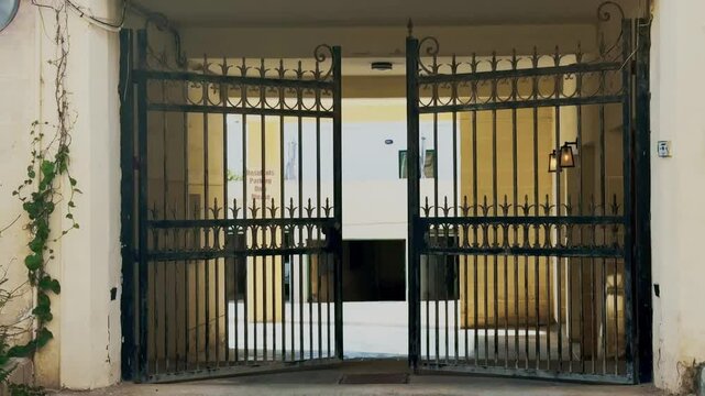 A black wrought iron gate slowly closes at the entrance to a residential parking area. The gate features decorative spikes and scrollwork. Warm light glows from wall lamps in the background.