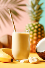 Tropical banana coconut pineapple smoothie in glass with striped straw on colorful summer background.