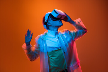 A person wearing VR goggles and vibrant attire explores a virtual reality setting under colorful lighting on an orange background.