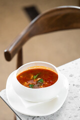 A bowl of tomato soup garnished with fresh cilantro, served on a white plate with a napkin. The dish is placed on a speckled countertop, with a blurred wooden chair in the background.