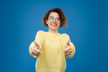 A happy young woman wearing glasses and a yellow sweater smiling with thumbs up. Vibrant blue...