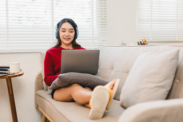 Fototapeta premium Young woman with long hair wearing headphones, red sweater, and slippers sits comfortably on sofa with pillow, using laptop and smiling in bright living room