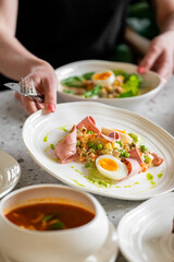 A plate with sliced ham, boiled egg, peas, and mixed vegetables being served. Another plate with similar food and a bowl of soup in the background. A close-up of a homemade meal presentation.