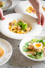 A close-up of a hand serving a plate of pasta with meatballs and fresh lettuce on a dining table, showcasing food presentation and culinary details.