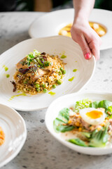 A hand serves a gourmet plate of risotto with peas, mushrooms, and microgreens on a table, surrounded by fresh and elegantly presented dishes in a bright setting.