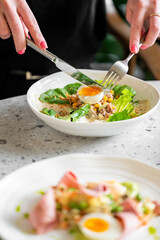 Close-up of a person slicing a soft-boiled egg on a fresh salad with lettuce, corn, and tuna. A second plate with a similar dish is blurred in the foreground, highlighting a healthy meal.