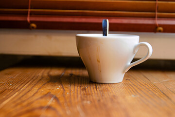 A white ceramic coffee cup with visible dried coffee stains and a metal spoon inside, placed on a rustic wooden windowsill in front of a window with closed wooden blinds. Soft focus
