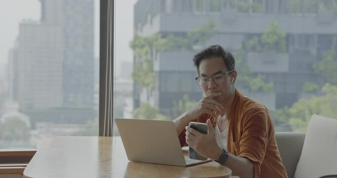 Senior Asian businessman wearing Bluetooth headset celebrates success during online meeting in modern co-working space. Remote work and business communication concept.