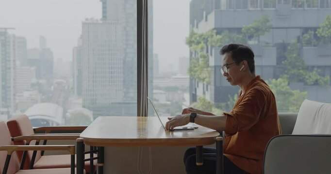Senior Asian businessman wearing Bluetooth headset celebrates success during online meeting in modern co-working space. Remote work and business communication concept.