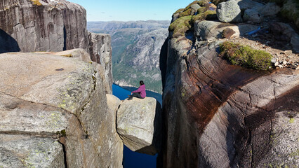 A lone adventurer sits atop a massive boulder, surrounded by steep cliffs and breathtaking fjords in Kjeragbolten Lysefjord, Norway. The blue sky enhances the serene atmosphere of this natural wonder.