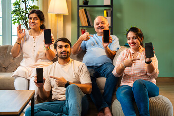 Happy Indian family seated on sofa indoors presenting smartphone screen with joyful expressions