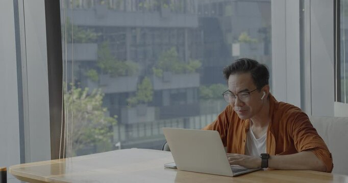 Senior Asian businessman wearing Bluetooth headset celebrates success during online meeting in modern co-working space. Remote work and business communication concept.