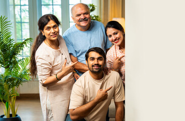 Coming behind board and smiling, Indian family of four showing blank white space with gestures