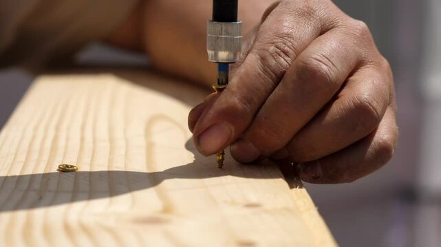 Man aligns two wood pieces, then uses a power drill to insert a long screw into the wooden frame. Detail on the drilling process.