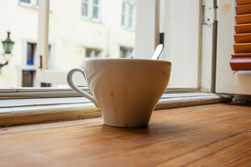 Closeup of a white ceramic coffee cup with a metal spoon, placed on a windowsill by an open window with a blurred old town of Tallinn in background. Soft focus 

