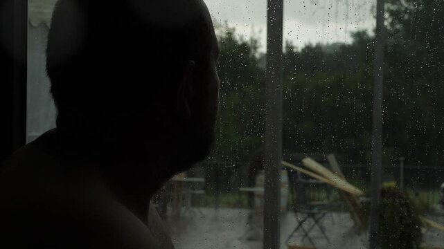 As the storm rages, a man looks out from behind a window, trapped in thought, like trying to recall a dream fading too fast.