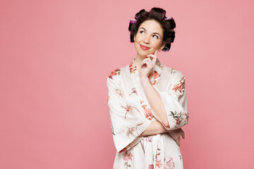 Young housewife housekeeper woman she wear white robe hair rollers put hand prop up on chin, lost in thought and conjectures isolated on pink color background studio portrait. Morning routine concept.