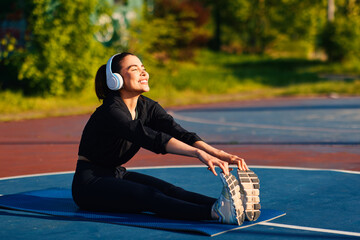 Happy athlete warming up and stretching legs on fitness mat, listening to music with headphones in outdoor workout area