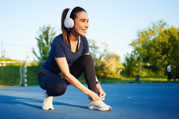 Young sporty woman tying shoelaces on her running shoes, preparing for outdoor workout, listening to music with wireless headphones