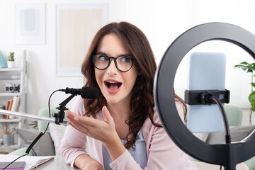 Smiling young female content creator and influencer at home, using a smartphone and ring light,...