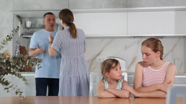 Frightened and frowning children sit at kitchen table during mom and dad quarrel. Behind children, parents swearing in background. Family conflict, divorce