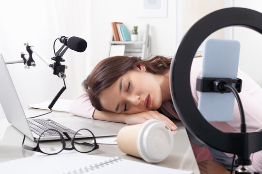 Exhausted female content creator and influencer asleep at desk with laptop, smartphone, and ring light. Tired and overworked from recording video tutorials, podcasts, or creating social media content