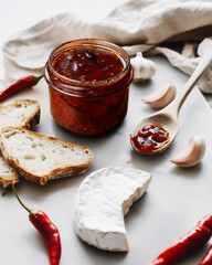 A jar of fiery red chili sauce on a light board, accompanied by Brie cheese wedge, toasted bread slices, garlic cloves, and a wooden spoon.