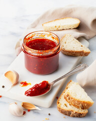 A vibrant jar of homemade red chili sauce surrounded by fresh garlic cloves and artisan bread slices on a light marble board.