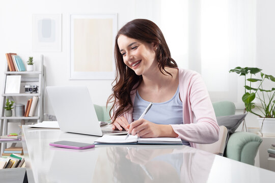 Happy young woman at home, sitting at a desk, using her laptop and taking notes with a pen in a notebook. Concept of positive remote work, online learning, or college studying with a joyful mindset