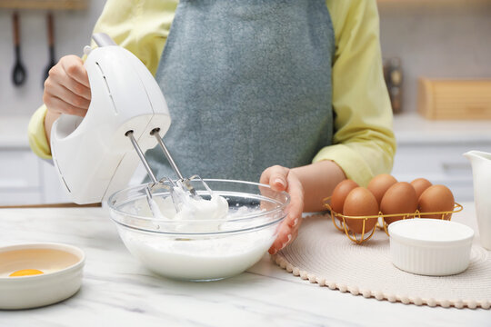 Woman whisking cream with hand mixer at white marble table indoors, closeup