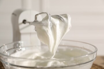 Hand mixer with whisks and bowl of whipped cream on blurred background, closeup