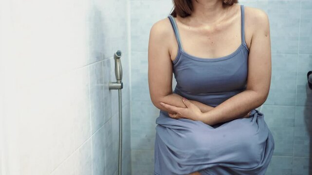 asian woman sitting on the toilet with stomach pain and cramps, suffering from diarrhea and bloating caused by ibs or chronic digestive issues