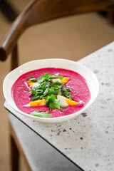A bowl of vibrant pink beet soup with chopped herbs and slices of boiled egg, placed on a speckled countertop. Fresh, colorful, and appetizing
